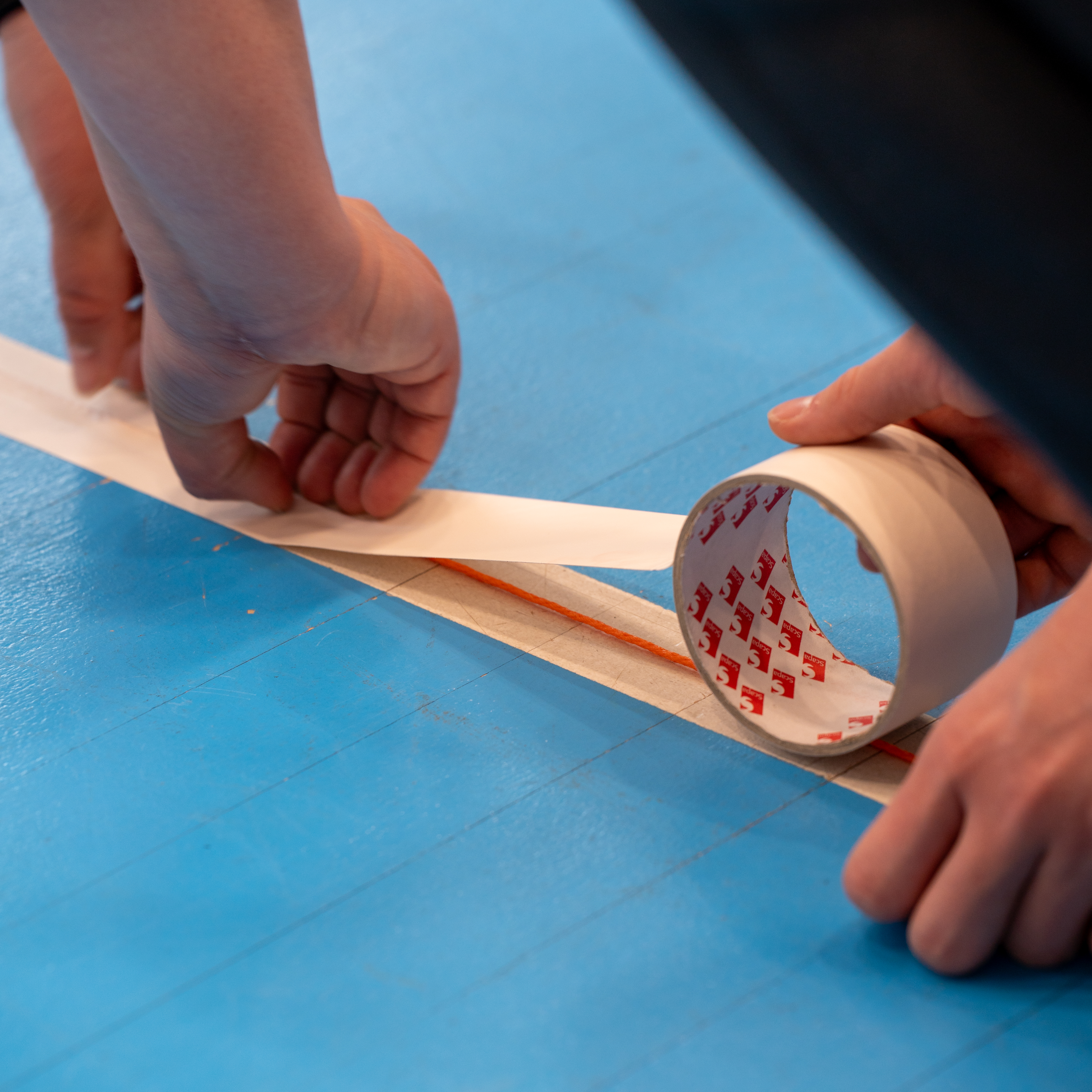 Kuvassa teipataan maalipallopelikentän viivojen alle narua, josta pelaajat tunnistaa missä he kentällä ovat.